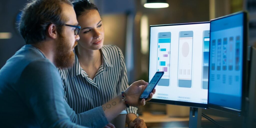 a male holding a smart phone and a female developer is seen infront of a monitor