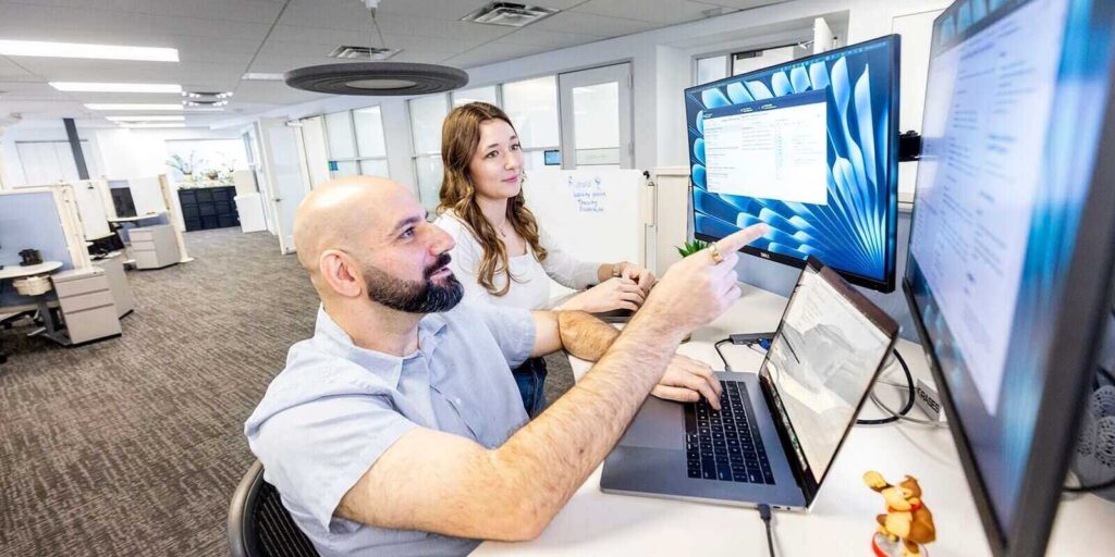 two colleagues collaborating on software development, analyzing code on dual monitors in a modern office environment
